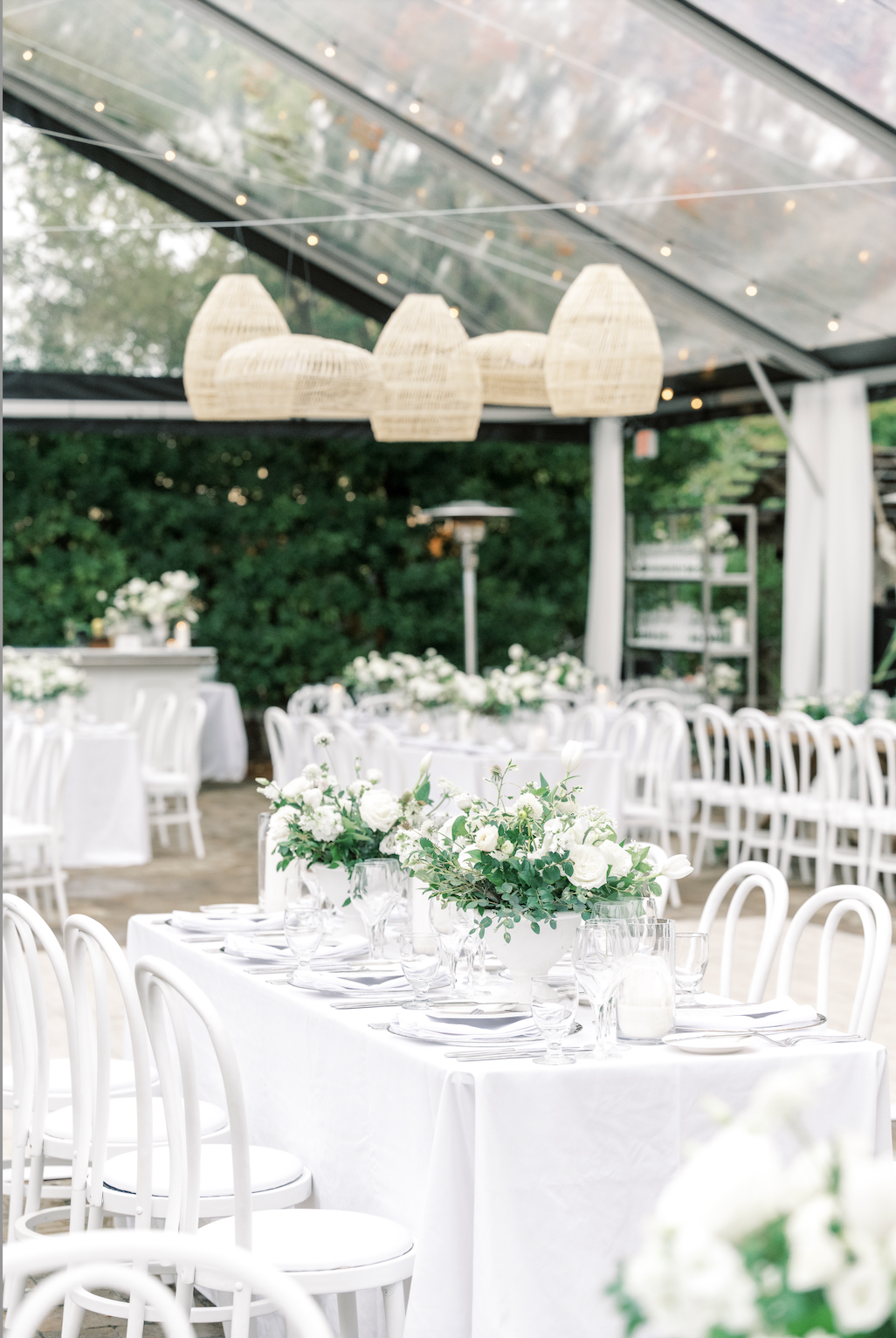 Elegant outdoor wedding setup with white chairs and tables under a clear tent. Tables decorated with white floral centerpieces; wicker pendant lights hang above.